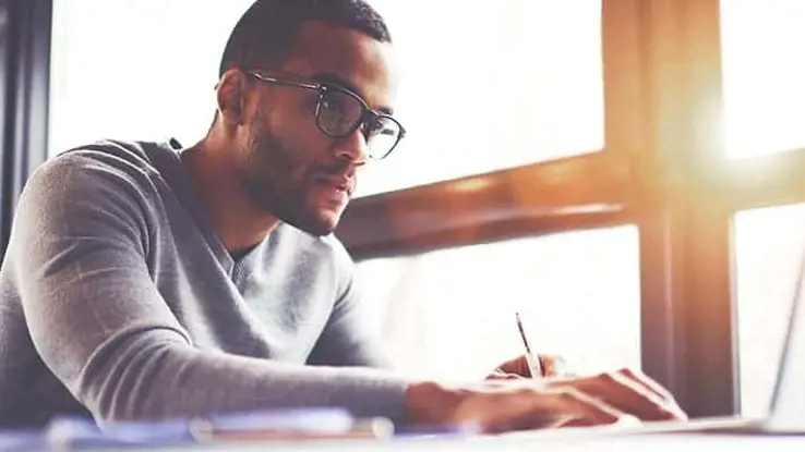 Side view of man with glasses looking intently at laptop while holding a pen