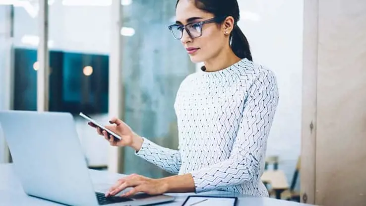 Side view of woman holding a phone in one hand and pressing on laptop keyboard with the other