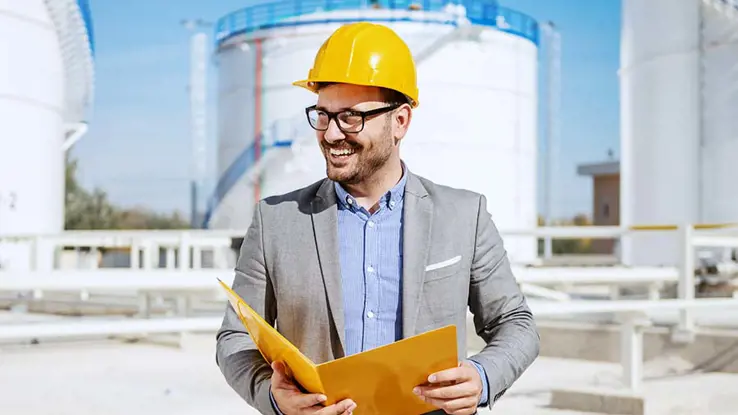 Smiling man in business suit and yellow hard hat holding an open folder at a construction site