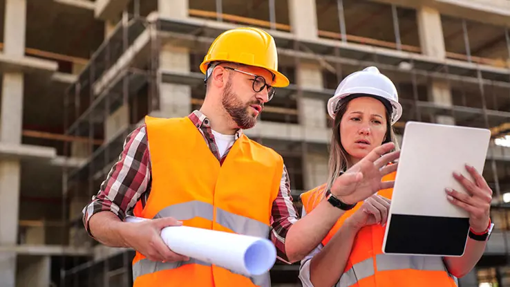 Male and female construction workers in hard hats and orange vests looking at tablet